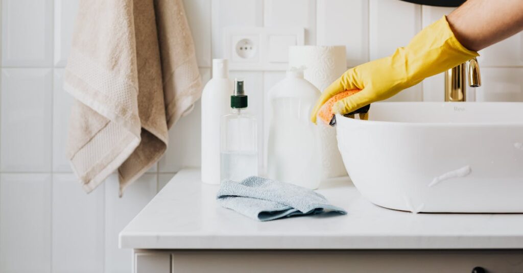 Person cleaning a bathroom sink with yellow gloves and cleaning products on a countertop.