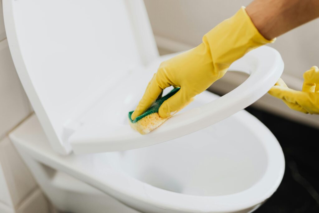 a person's hand with a yellow rubber glove is holding a yellow cleaning sponge and cleaning the toilet seat and the toilet