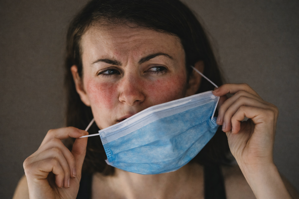 Woman taking off a face mask with red, irritated skin around the nose and mouth, illustrating mask-related reactions in sensitive individuals.